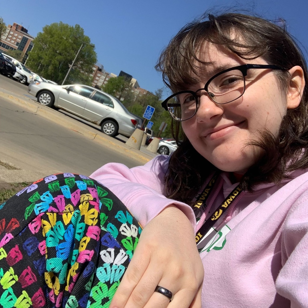 A picture of Vera. They are sitting in a parking lot and are smiling. They have a pink sweatshirt on as well as colourful pants.