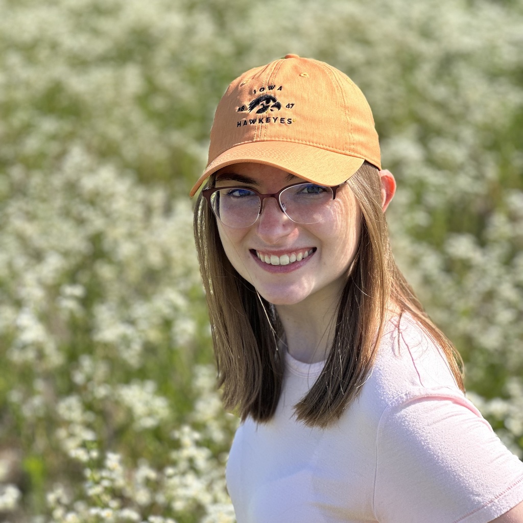 A picture of Tori. She is standing in a field and smiling. She is wearing a white tank top and a yellow baseball cap.