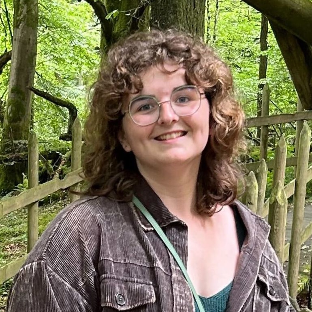 A picture of Olivia. She is standing in a forest and smiling. She is wearing a green tank top and a dark denim jacket.