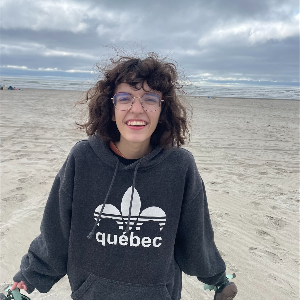 A picture of Maia. She is on the beach and is smiling. She is wearing a dark grey sweatshirt that says "Quebec" and is holding her shoes in her hands.