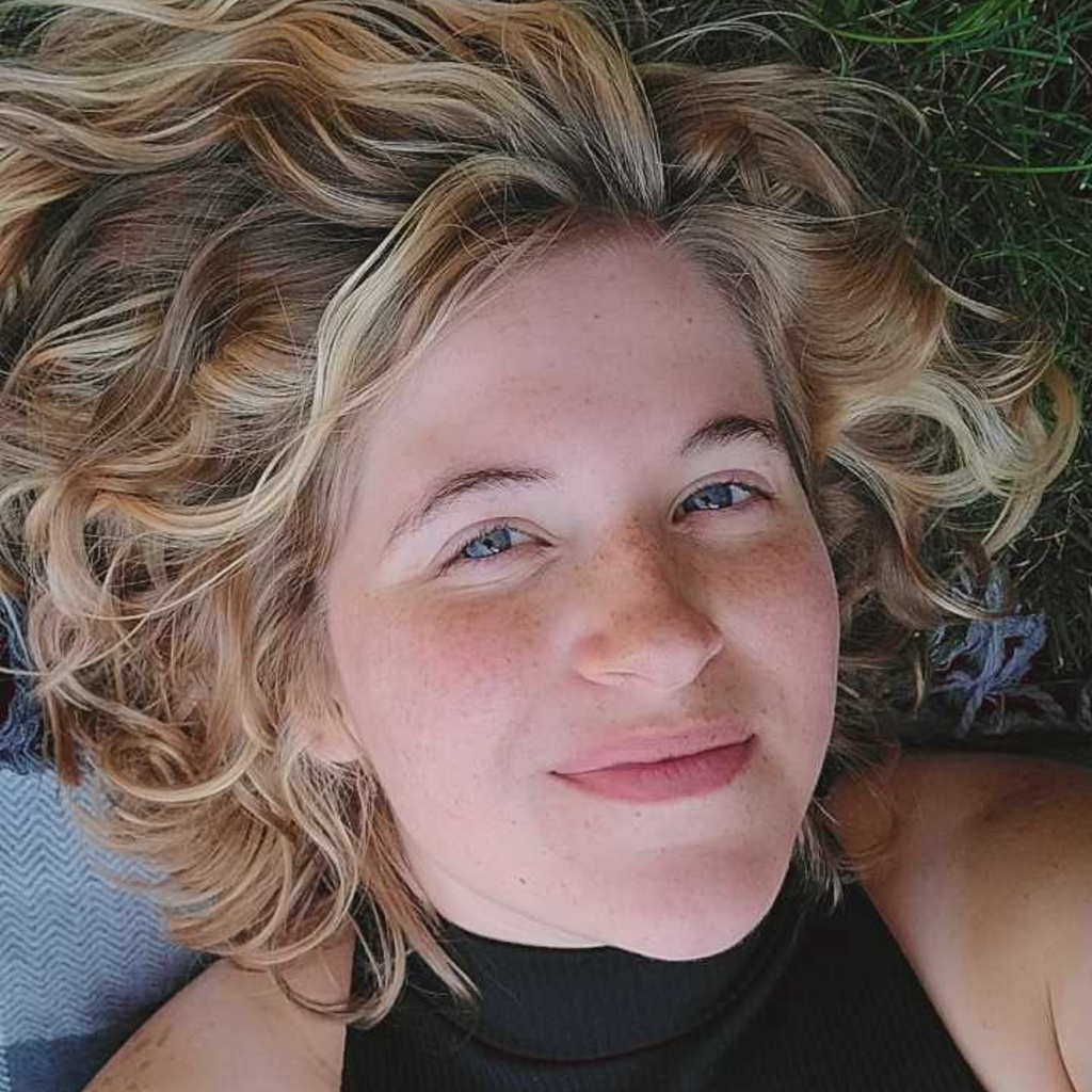 A picture of Dennie laying on a picnic blanket in the grass. Their hair is fanned out behind them as they smile at the camera.