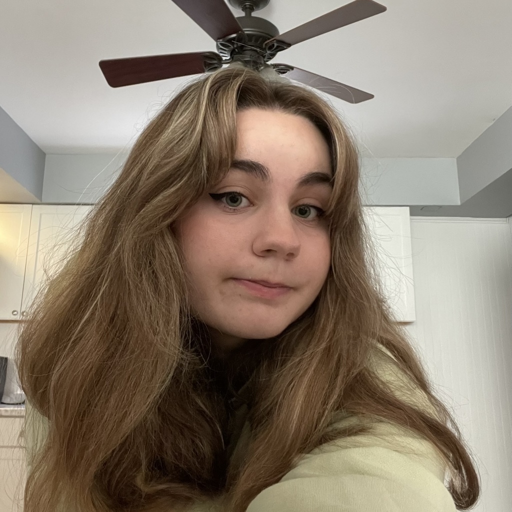 A picture of Emma with her hair down, holding the camera out in front of her and smiling. In the background is a ceiling with a bulkhead and a fan.