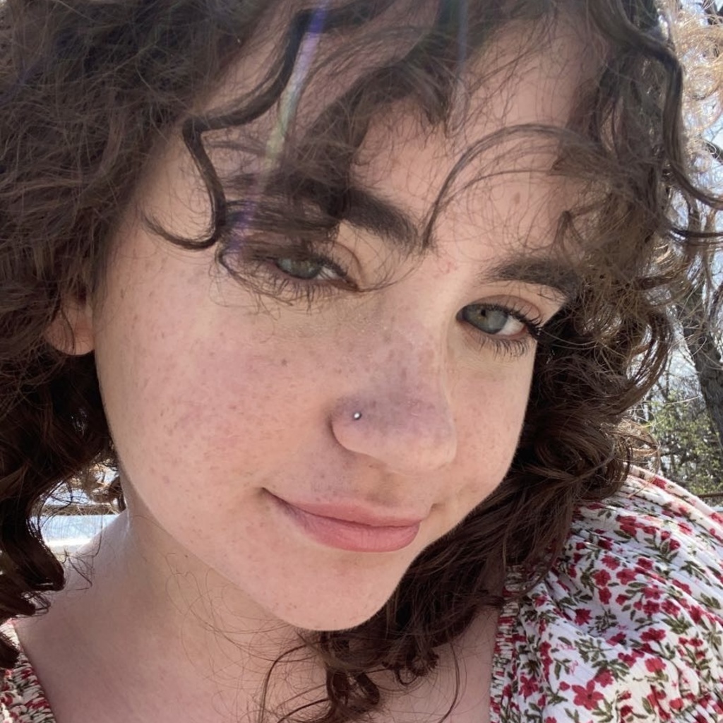 A picture of Rachel. Her curly hair falls to her shoulders, bangs sweeping across her forehead. Her head is tilted to the side as she smiles softly.